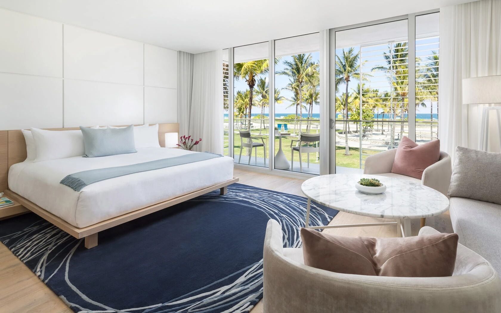 A hotel room with a bed, a round table, and a view of palm trees and the ocean.