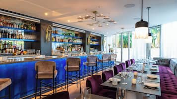 Un espace bar doté d’un comptoir bleu, de plusieurs tabourets de bar et d’un coin salon avec des fauteuils et des tables recouverts d’un tissu violet.