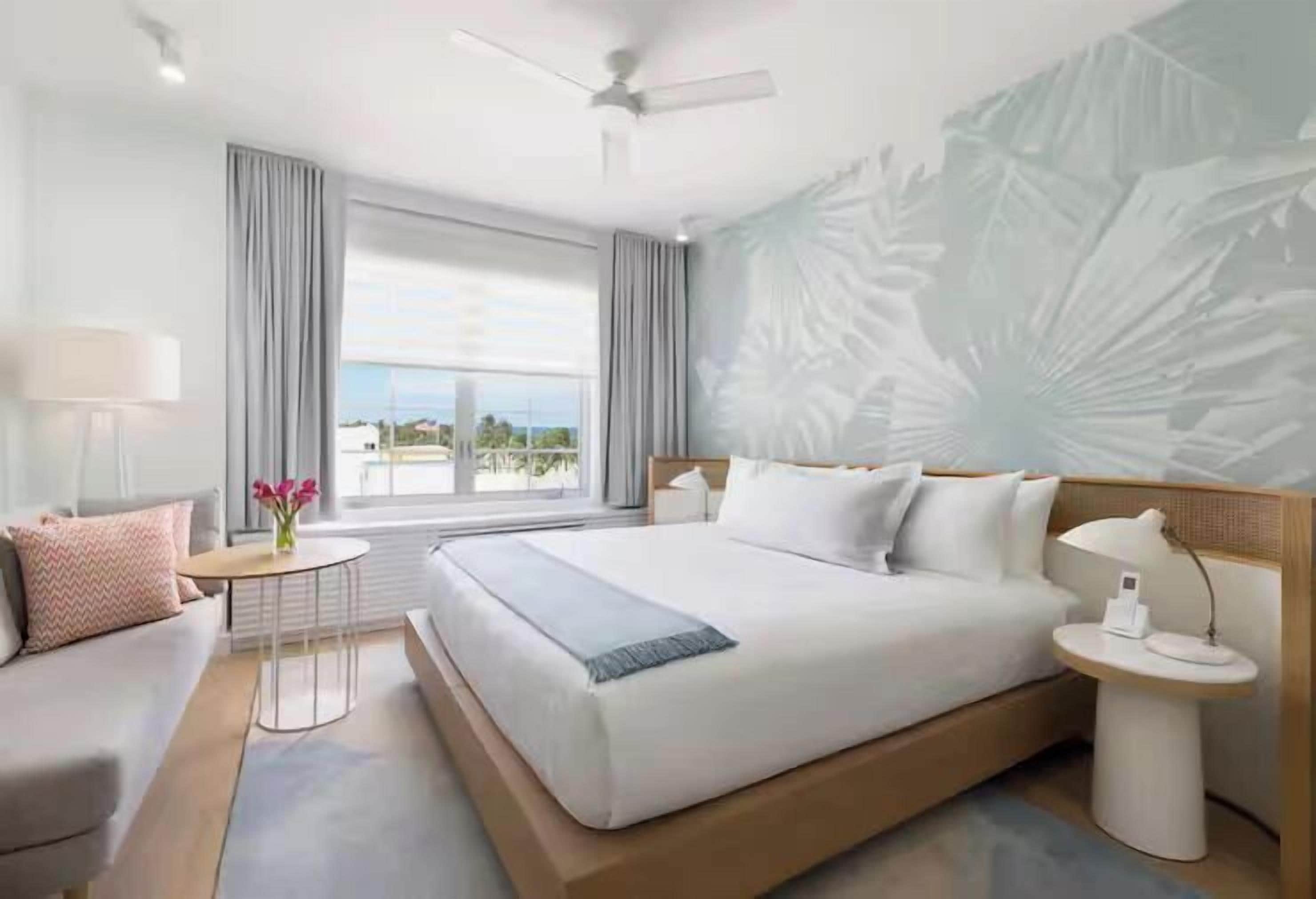 A modern hotel room with a large bed, a small table, a nightstand, and a view of the outdoors.
