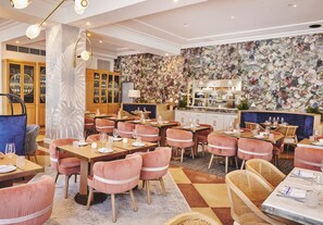 Un restaurant avec des tables et des chaises en bois, un mur décoré d’une fresque florale et un bar avec des étagères remplies de bouteilles.