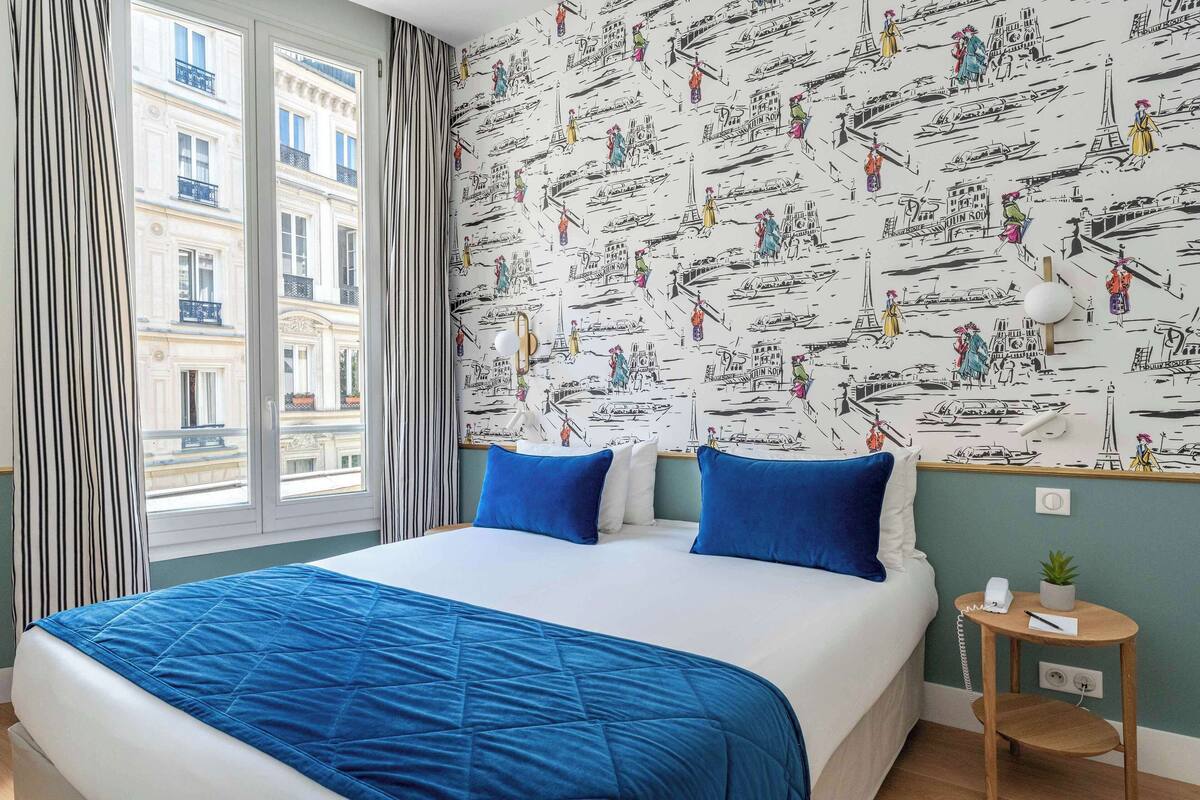 Habitación de hotel con una cama grande, ropa de cama azul, una ventana con cortinas, un papel tapiz de temática náutica en la pared y una mesita de noche con un teléfono y una planta.