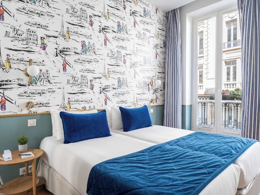Habitación de hotel con una cama grande, dos almohadas, una mesita de noche con lámpara, una planta pequeña y una ventana con cortinas.