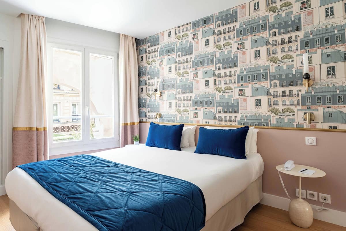 Una habitación de hotel con una cama grande, una mesita de noche con lámpara y una pared con papel tapiz de paisaje urbano.