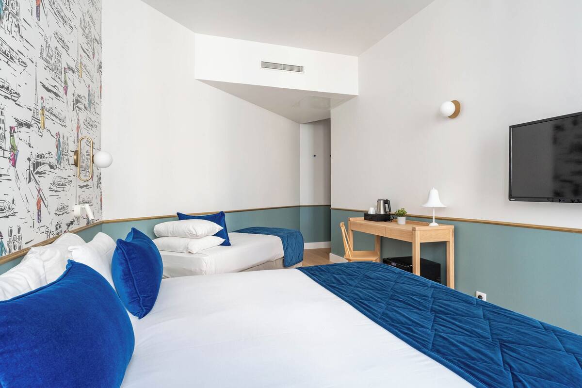 Habitación de hotel con dos camas, un escritorio, una silla, un televisor y una pared con papel tapiz de paisaje urbano.