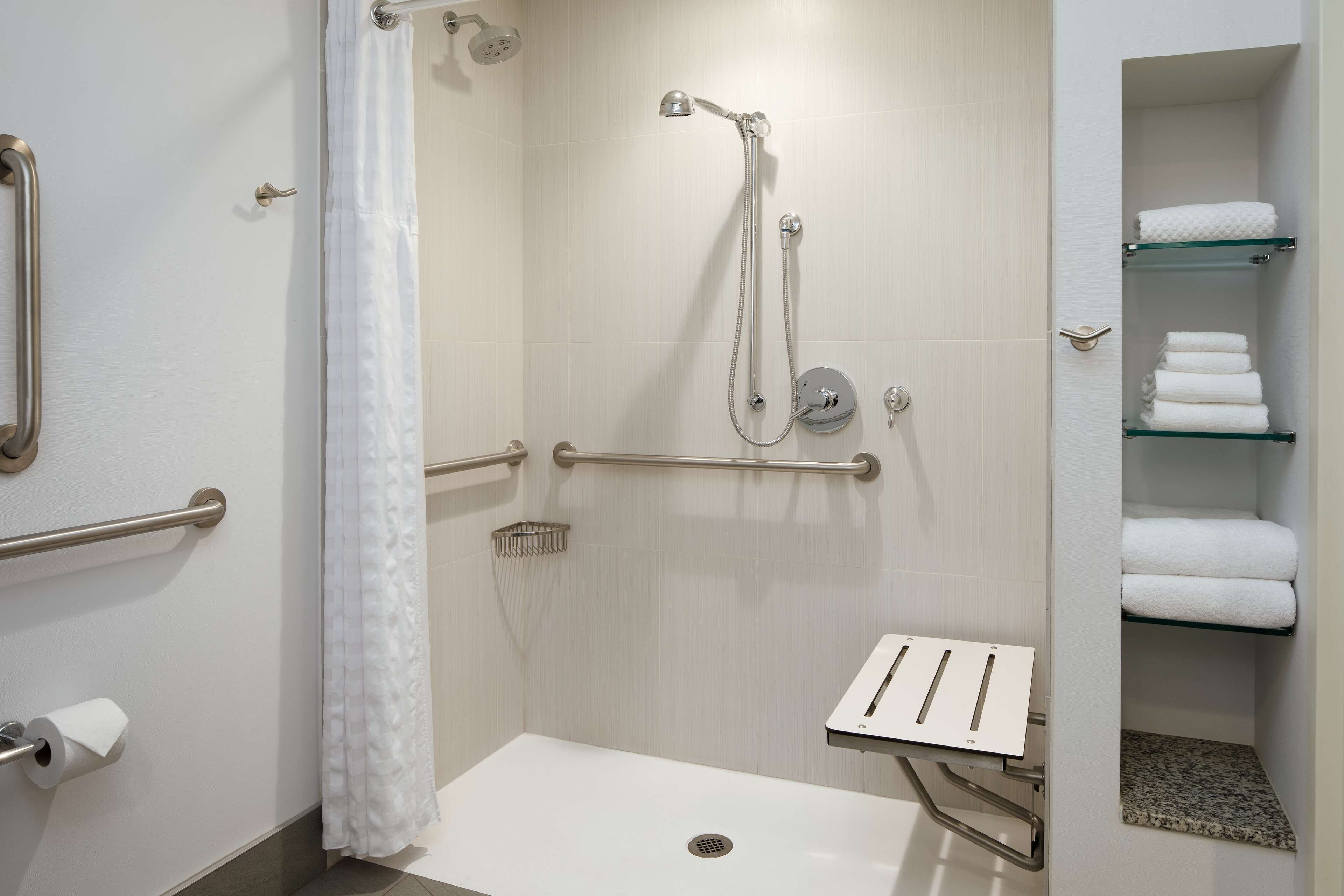 A modern bathroom with a walk-in shower, grab bars, a glass shelf with towels, and a toilet paper holder.