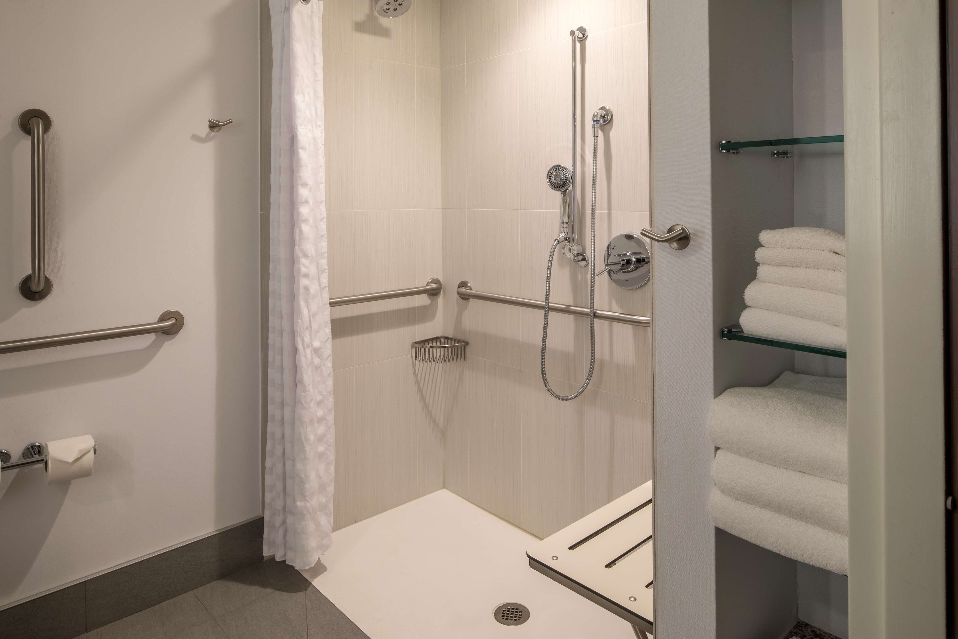 Un baño moderno con ducha a nivel, barras de apoyo, un estante de vidrio con toallas y un dispensador de papel higiénico.