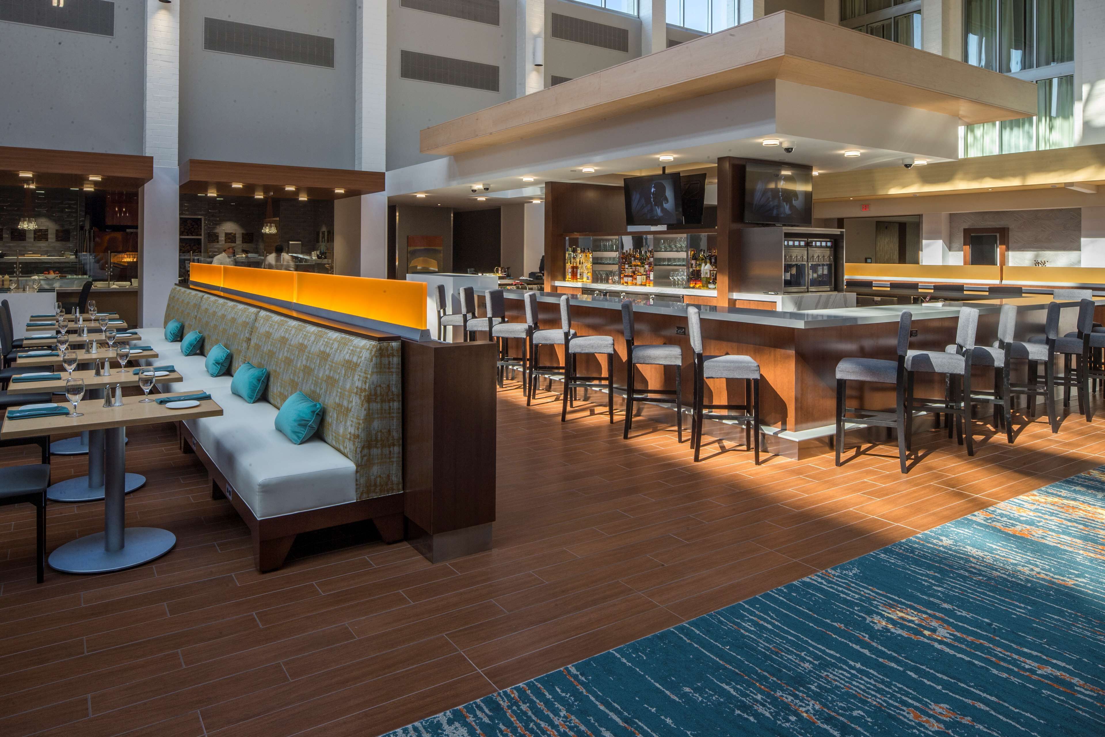 A modern hotel lobby with a bar area, seating arrangements, and a view of the kitchen.