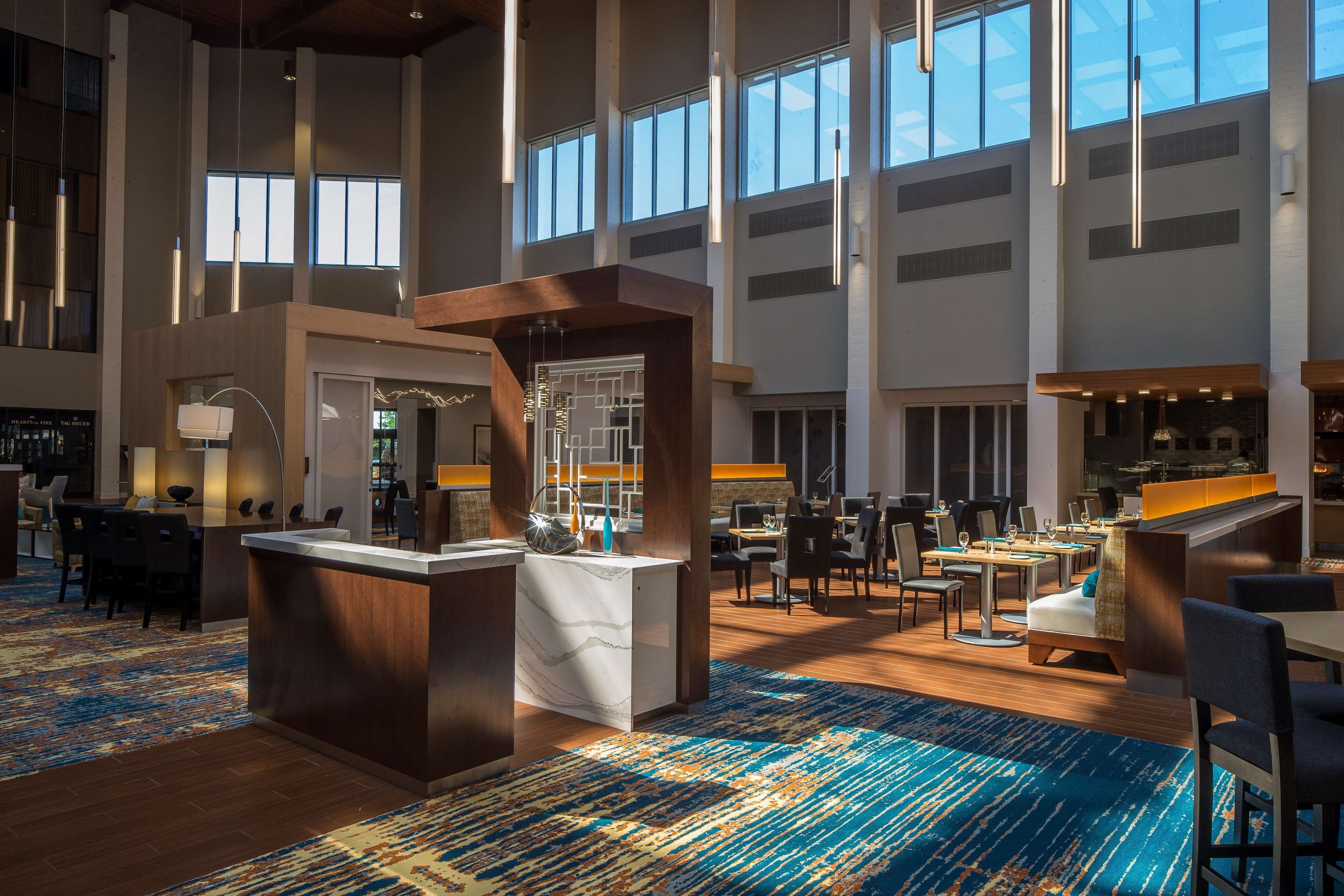 A spacious hotel lobby with high ceilings, large windows, a central reception desk, multiple dining areas, and a patterned carpet.