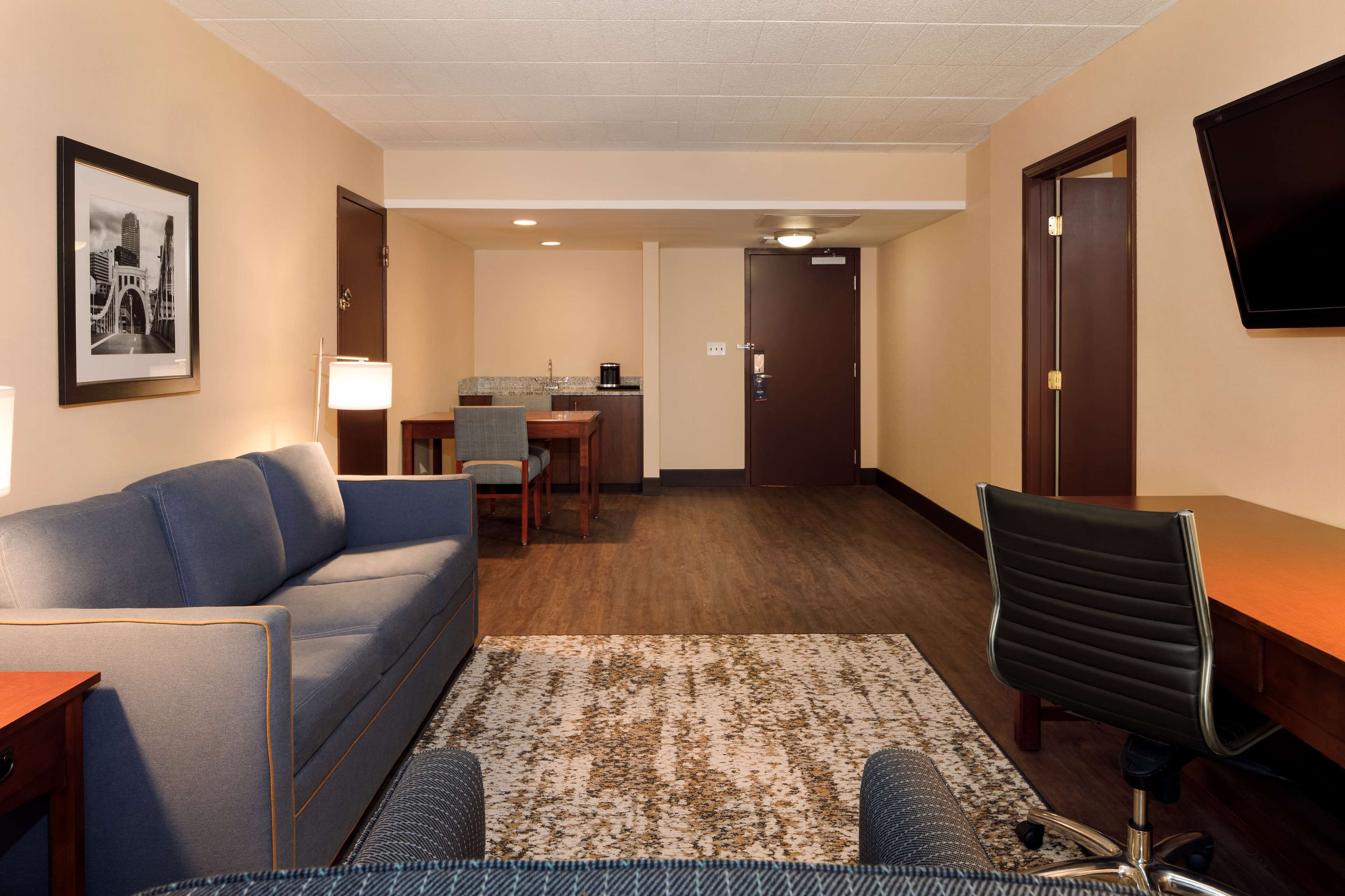 A hotel room with a sofa, a desk, a chair, a small kitchen area, and a television mounted on the wall.