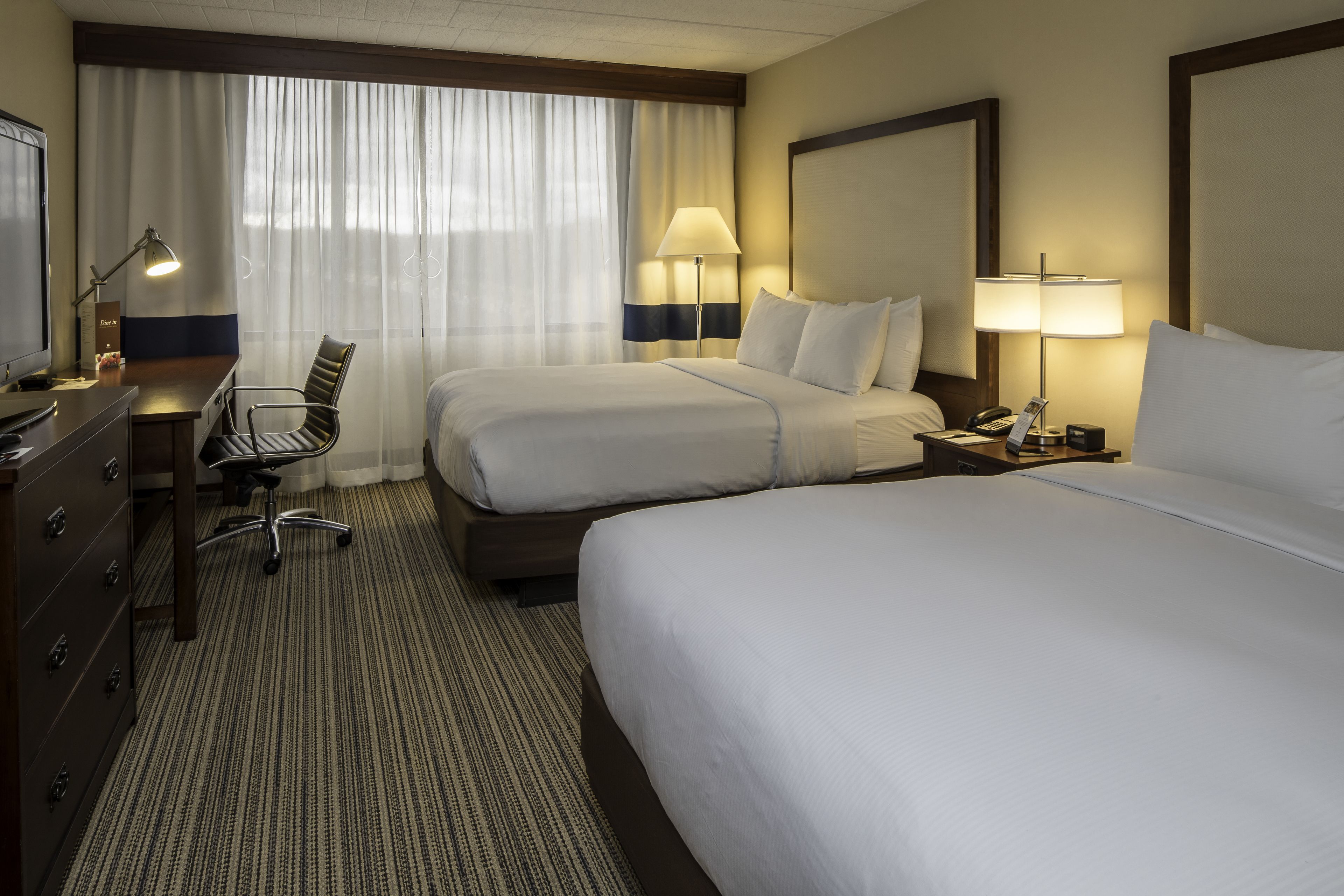 A hotel room with two beds, a desk with a chair, a dresser, and two lamps.