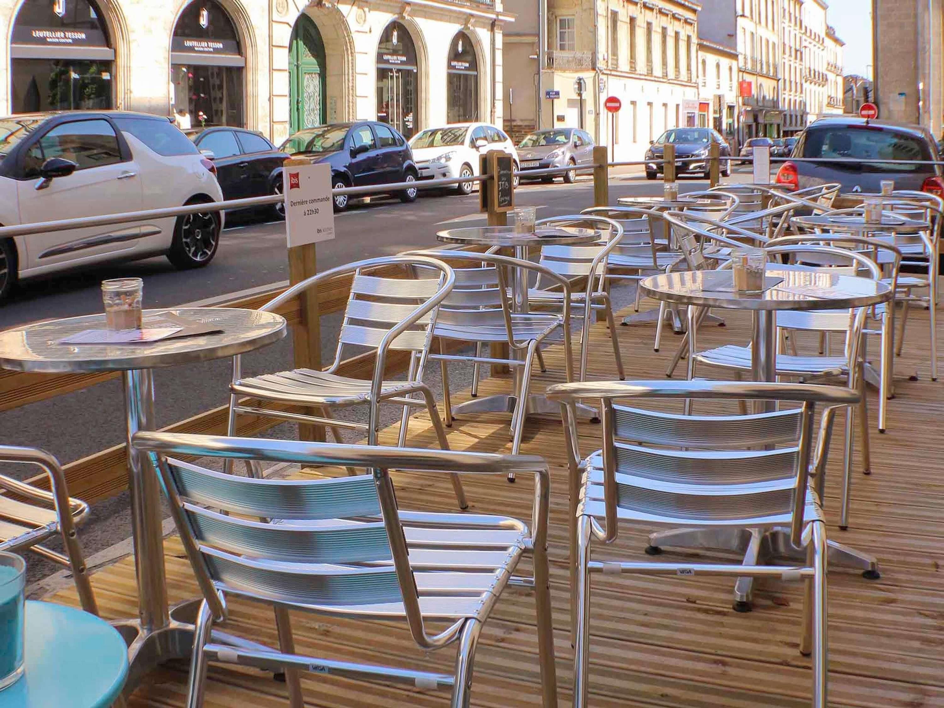 Café mit Außenbestuhlung, Tische und Stühle, eine Straße mit parkenden Autos und Gebäude im Hintergrund.