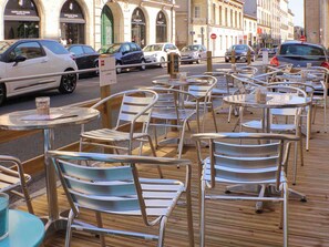 Terrasse de café avec tables et chaises, une rue bordée de voitures garées et des bâtiments en arrière-plan.