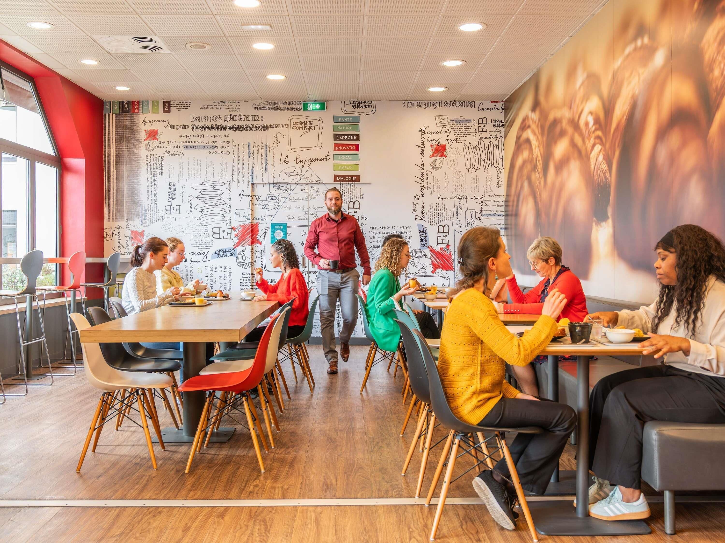 Un espace de restauration moderne avec des tables et des chaises en bois, une grande fresque murale et une personne qui traverse l’espace.