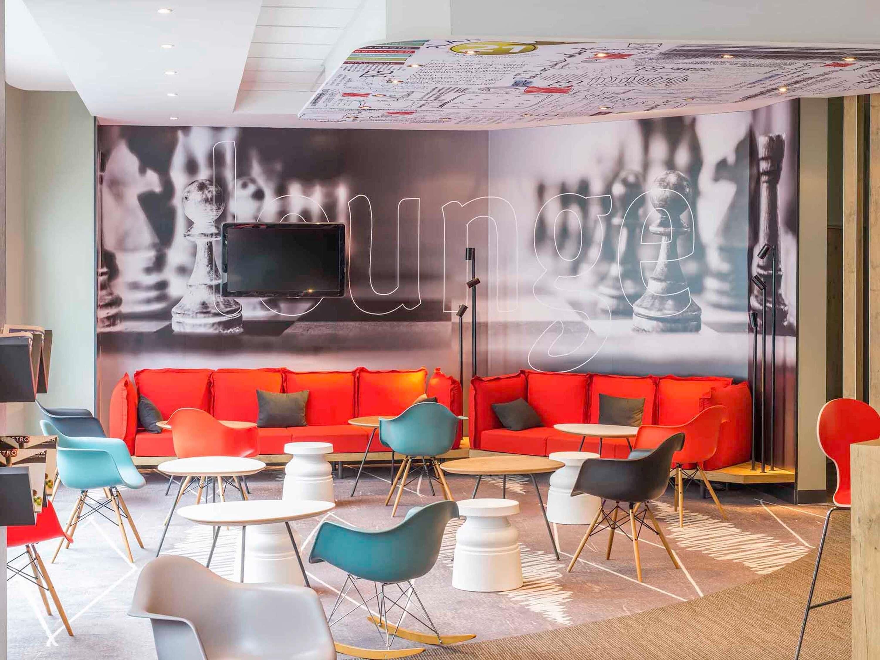A modern lounge area with a red sofa, teal and black chairs, and a large wall-mounted TV.