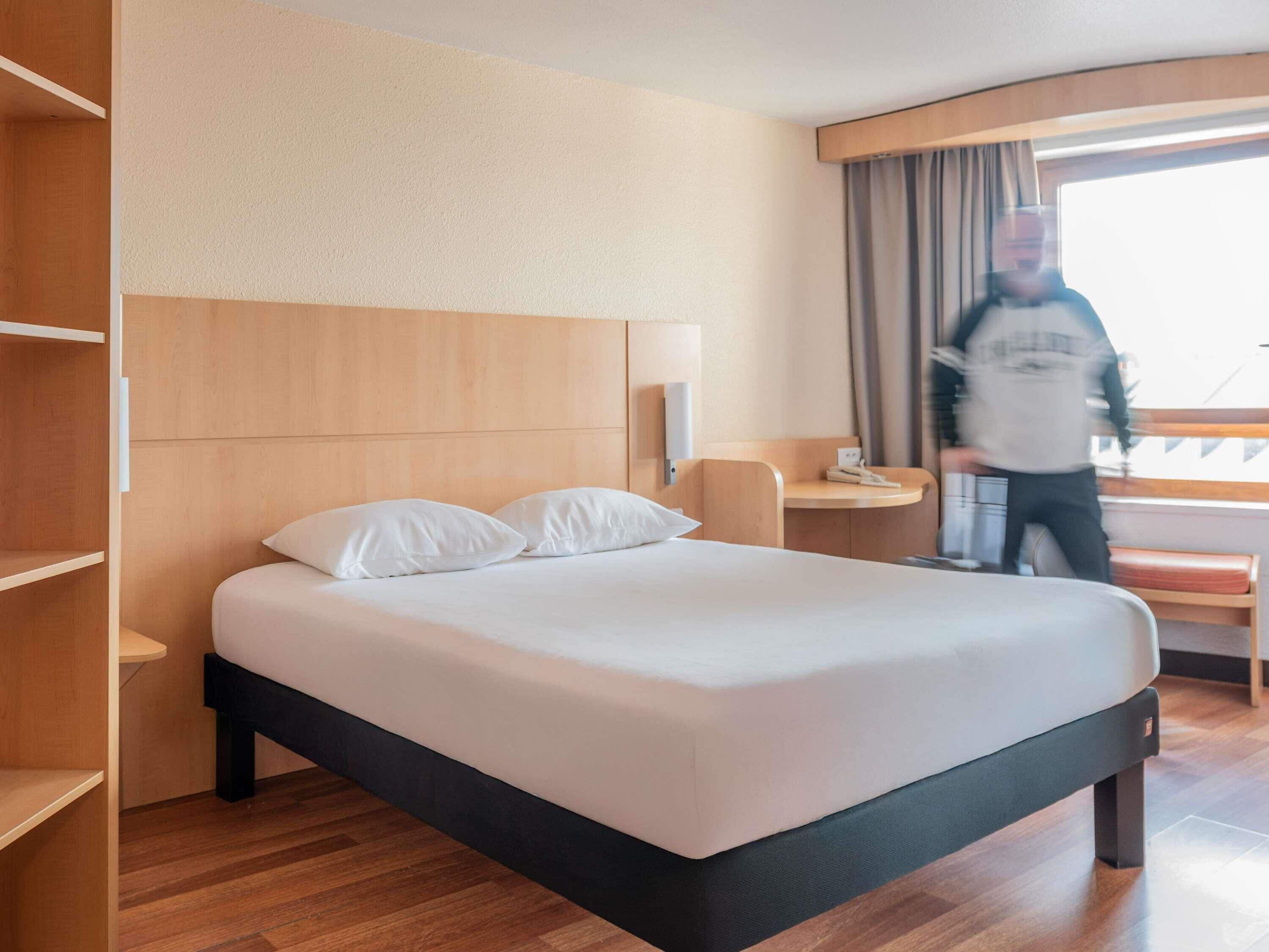 A hotel room with a bed, a bedside table, a lamp, a chair, and a window with curtains.