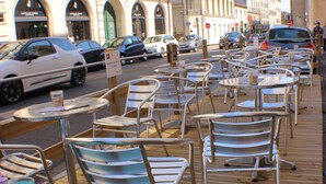 Une terrasse en bois, avec des chaises et des tables en métal, donnant sur une rue bordée de voitures garées et d’immeubles.