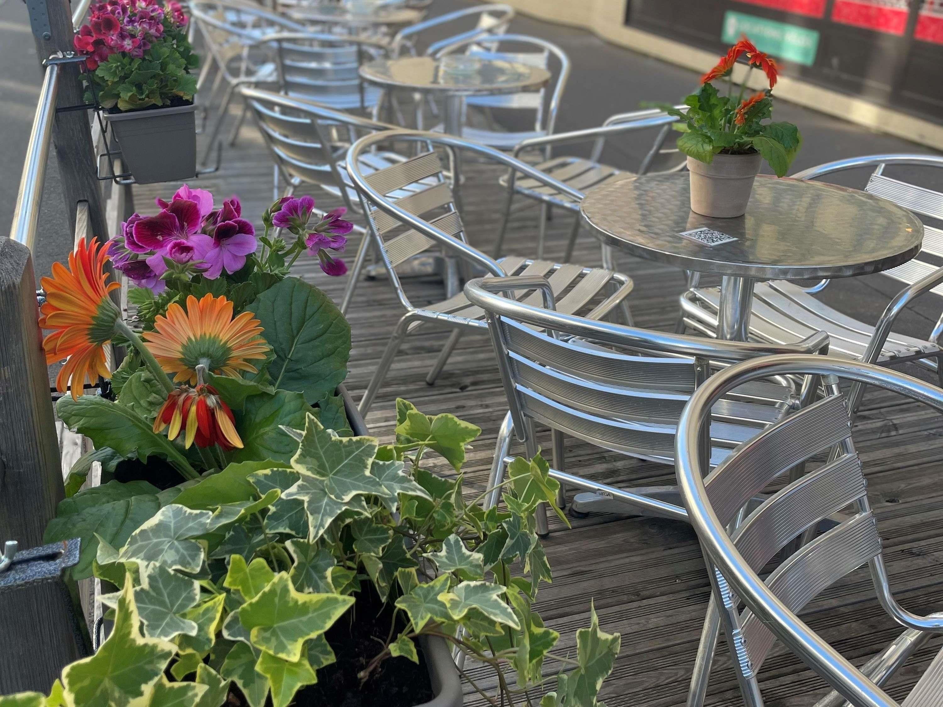Outdoor seating with metal chairs and tables, potted plants with flowers, and a wooden deck.