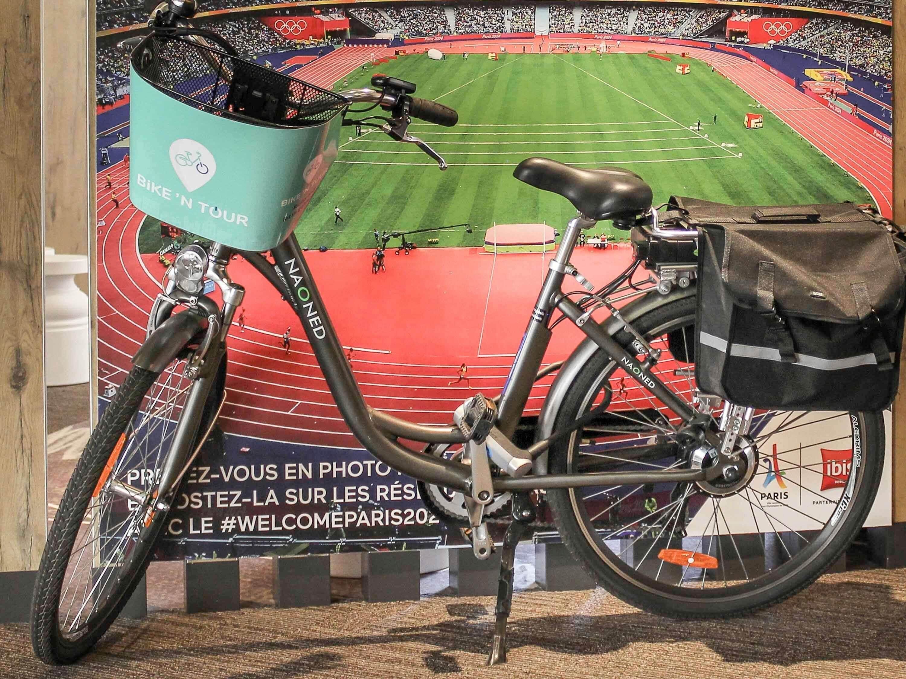 Een fiets met een mandje en tas, geparkeerd voor een stadion met een 'Welcome Paris 2022' bord.