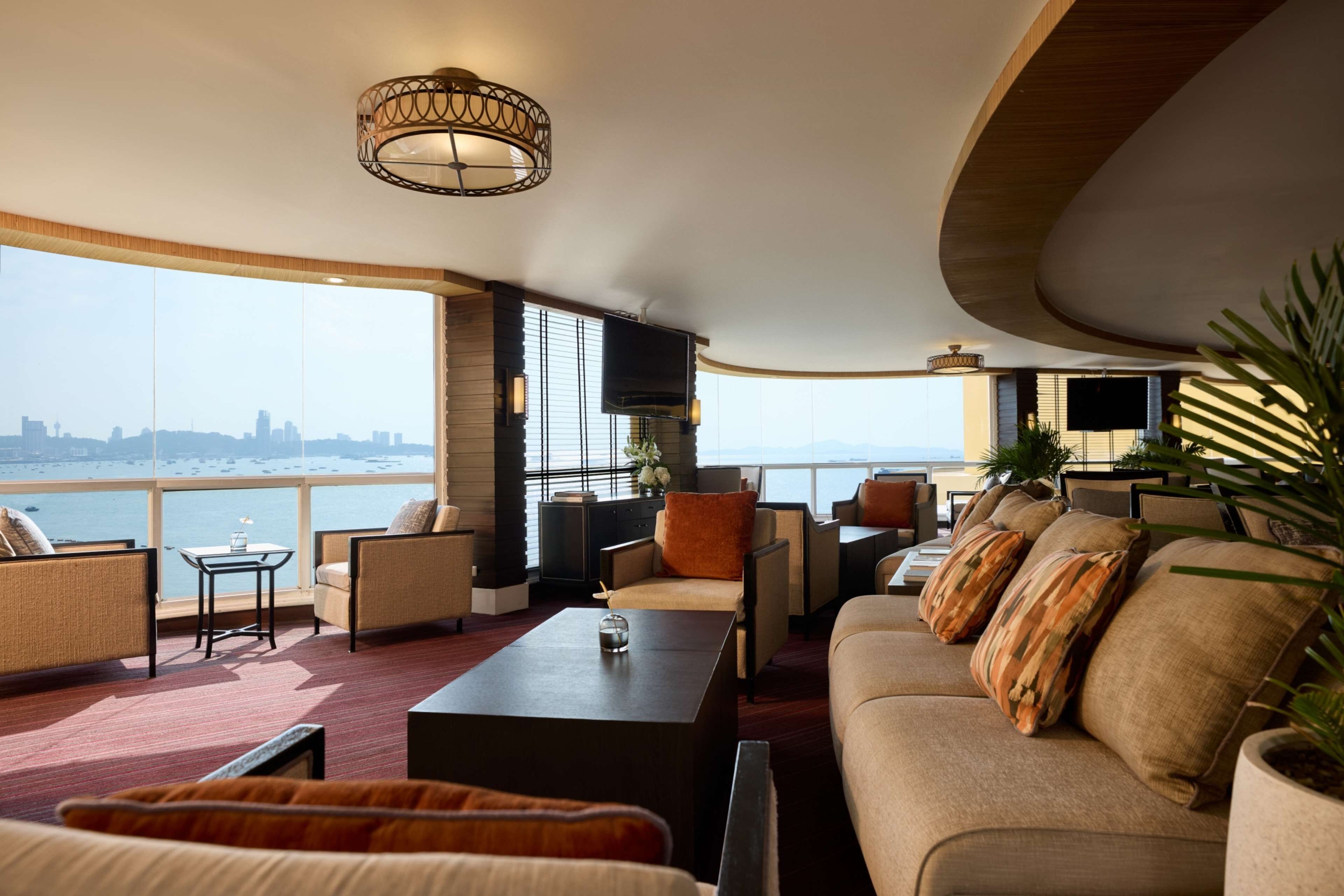 A spacious living area with a large sofa, a coffee table, and a TV mounted on the wall. There are multiple seating options, including a sofa and chairs. The room features large windows with a view of the city skyline and the sea.