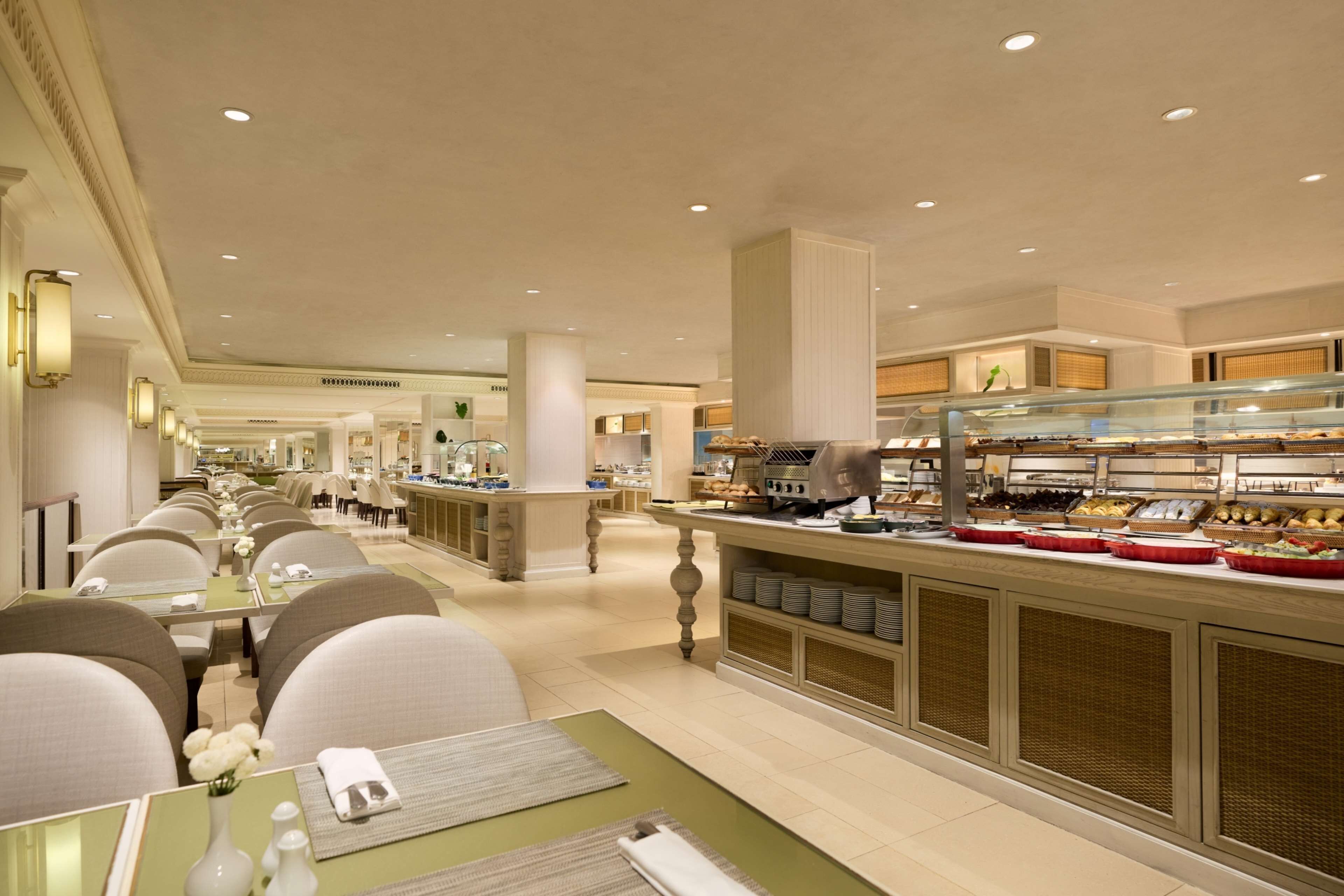 A hotel dining area with multiple tables and chairs, a buffet counter with various food items, and a clean, well-lit interior.