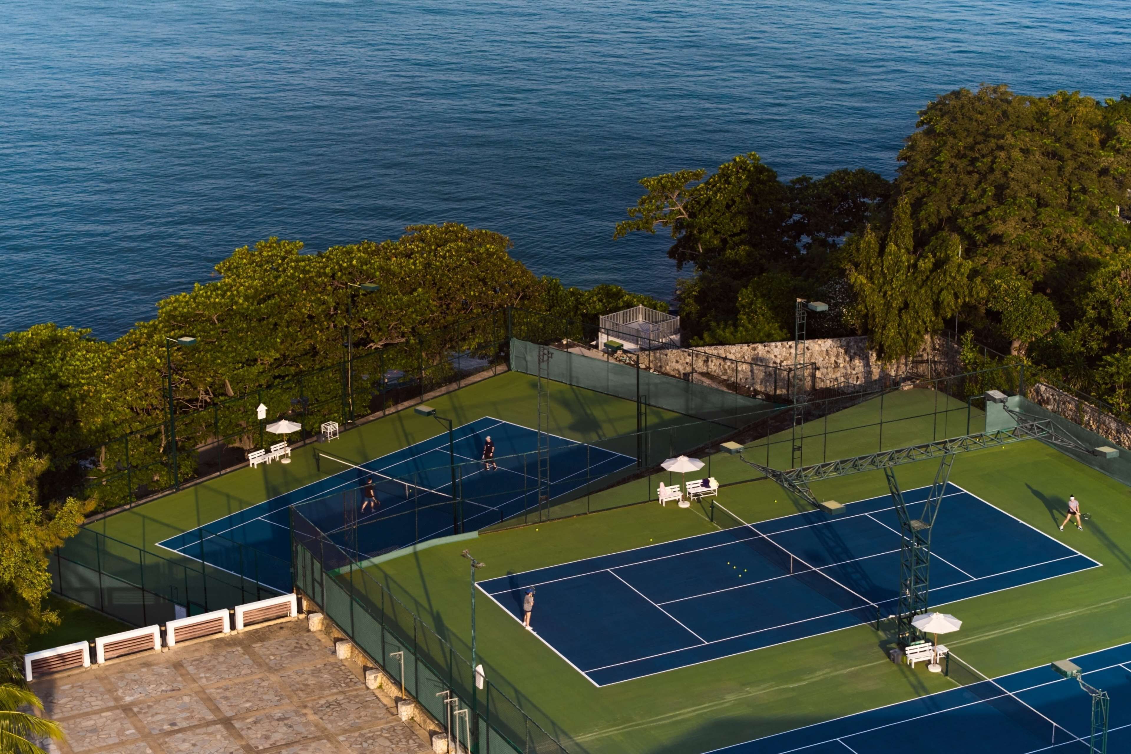 An aerial view of a tennis court complex with multiple courts, surrounded by trees and overlooking a body of water.