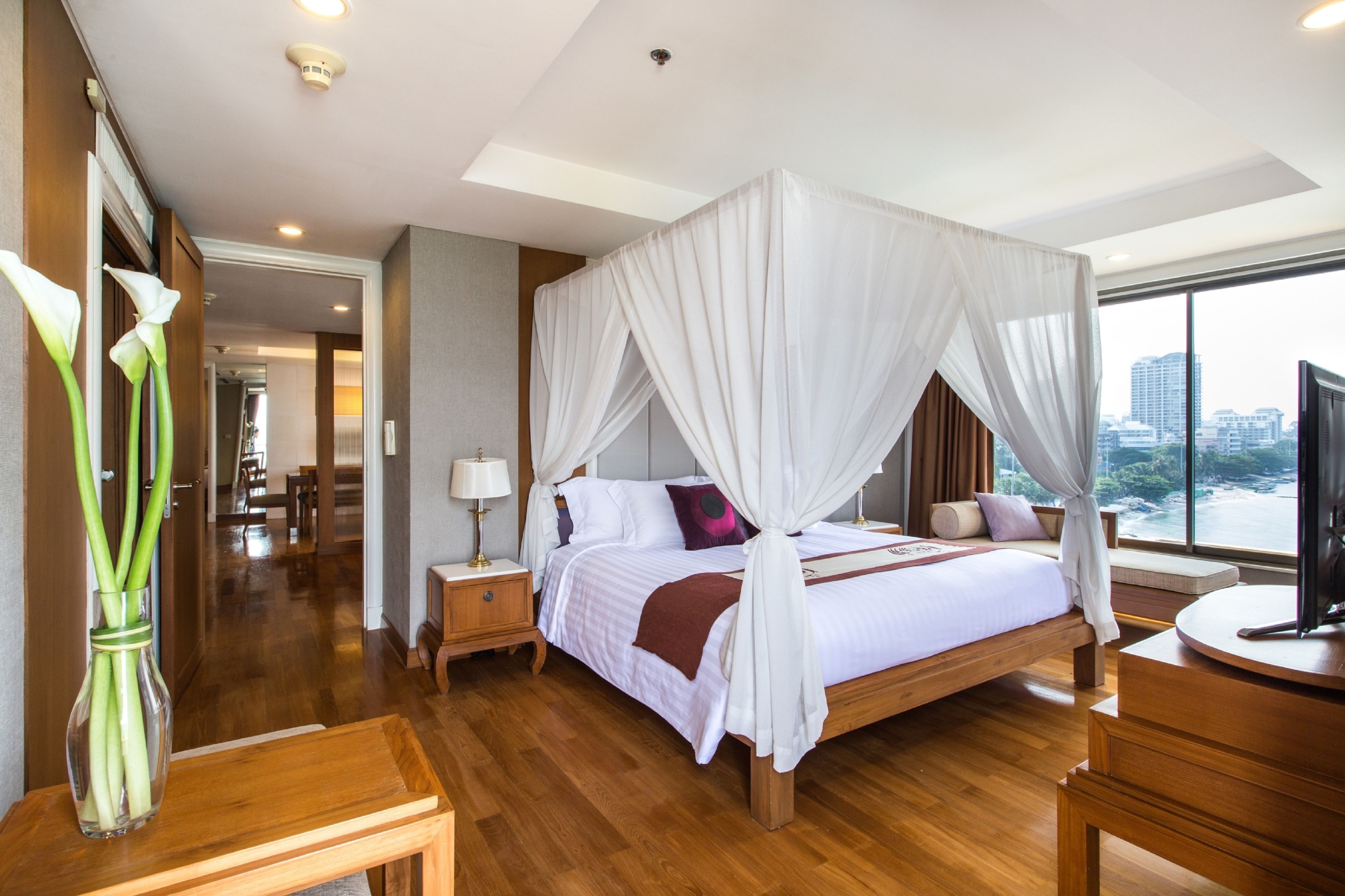 A spacious bedroom with a canopy bed, wooden flooring, a bedside table with a lamp, and a view of the city through large windows.