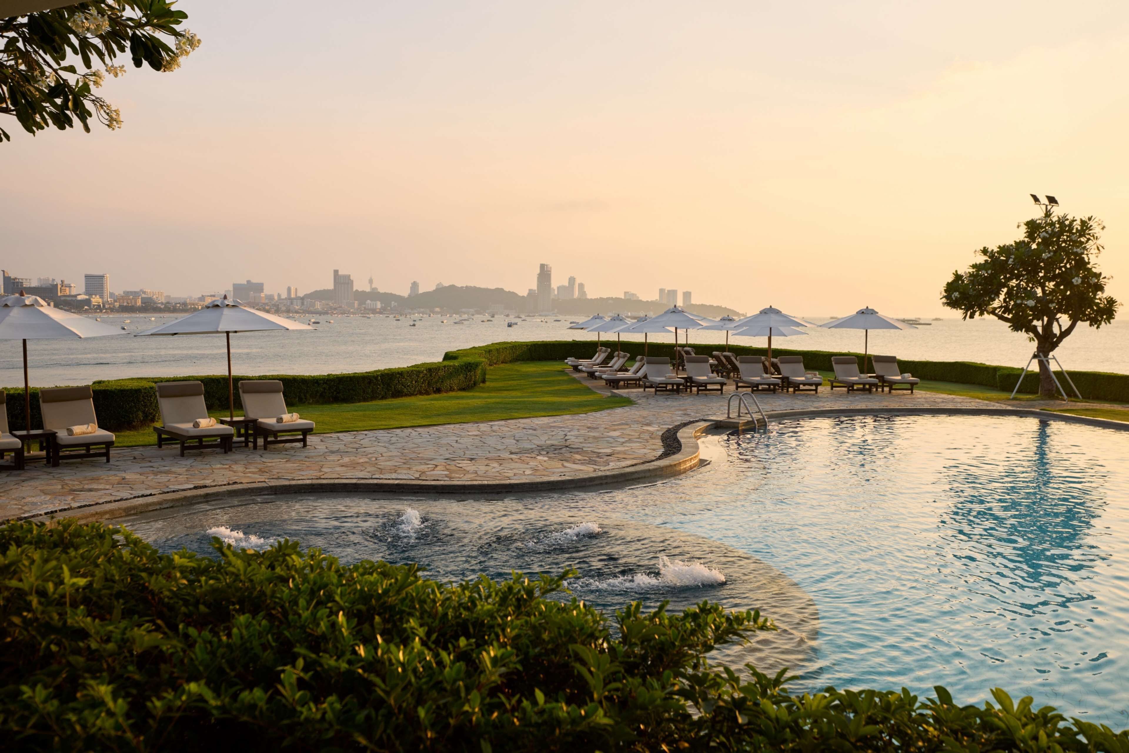 A pool area with lounge chairs and umbrellas, surrounded by greenery and a view of the city skyline.