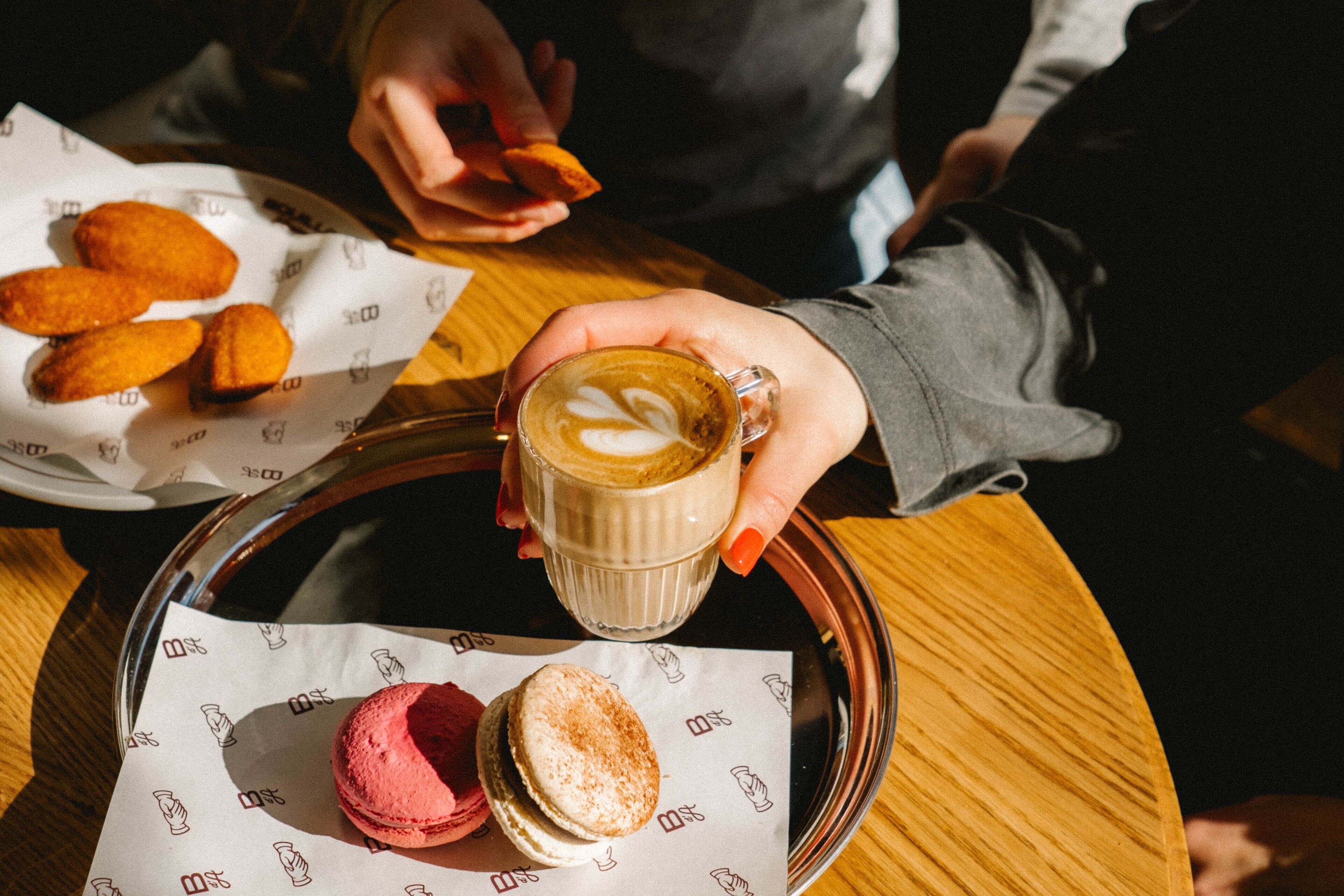 Una persona sosteniendo una taza de café con un diseño de arte latte, una bandeja con macarons y croissants, y una mesa con un motivo de logo.