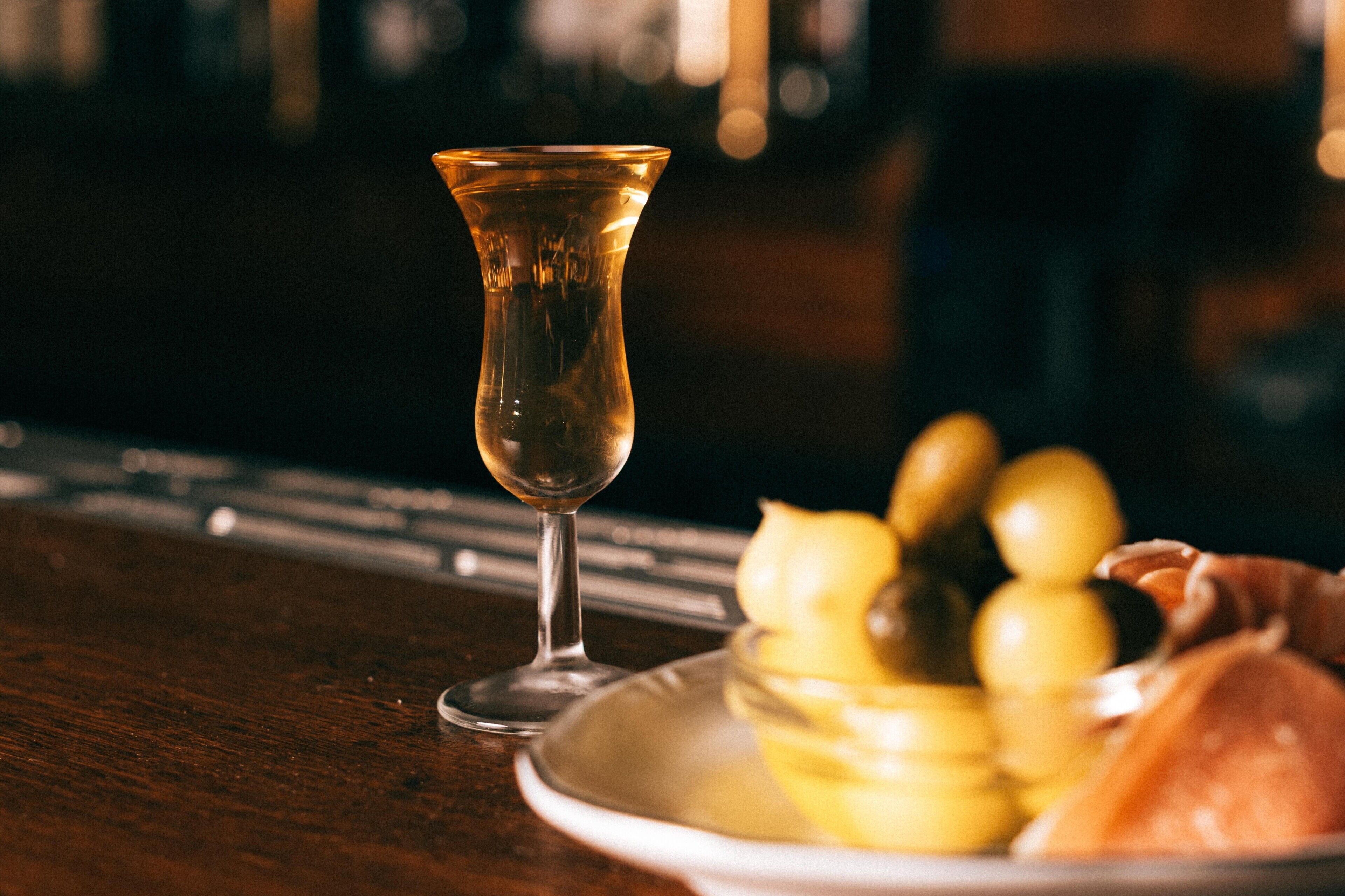 A glass of a clear beverage on a wooden surface with a plate of olives and a piece of bread.
