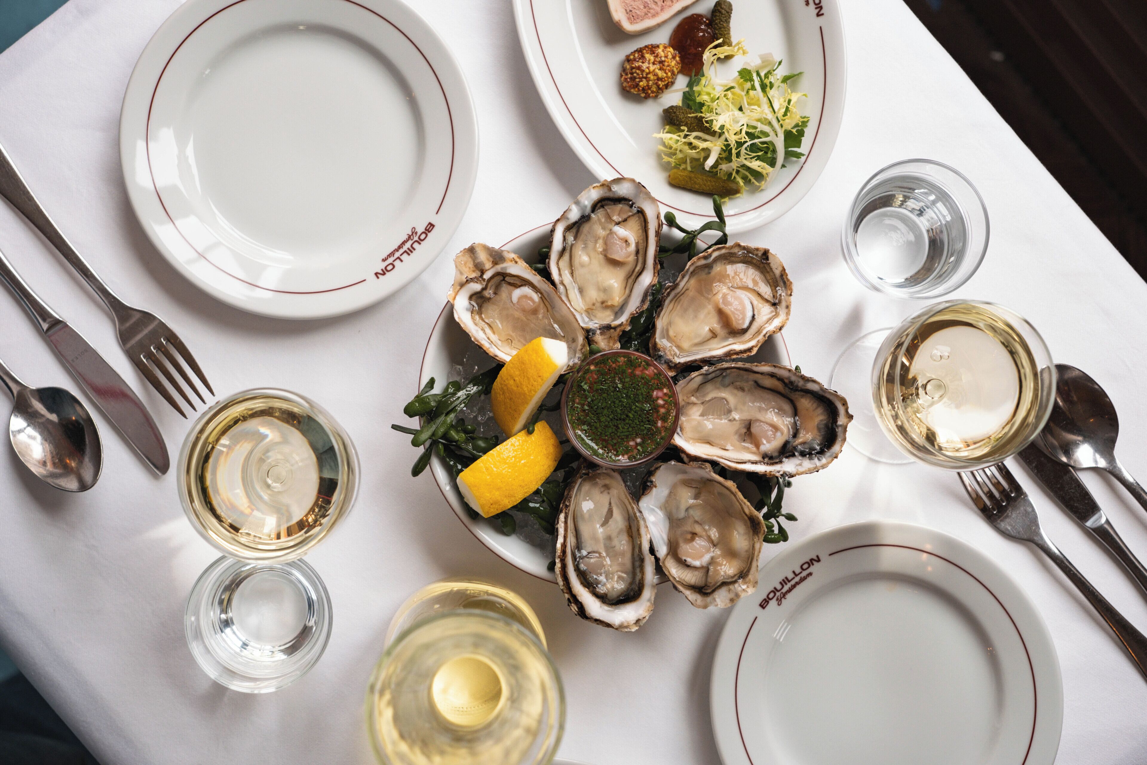 A dining table with plates of oysters, glasses of white wine, and a salad.