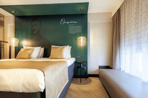 Une chambre d’hôtel avec un lit, une petite table, un banc, un mur orné d’une fresque et un rideau à motifs.