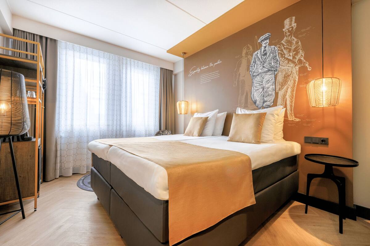 Una habitación de hotel moderna con una cama grande, un escritorio con silla, una lámpara de pie y un aplique de pared.