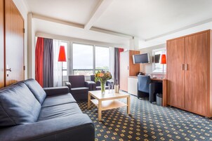 A hotel room with a sofa, a small table with a vase and glasses, a desk with a computer, and a TV on the wall.