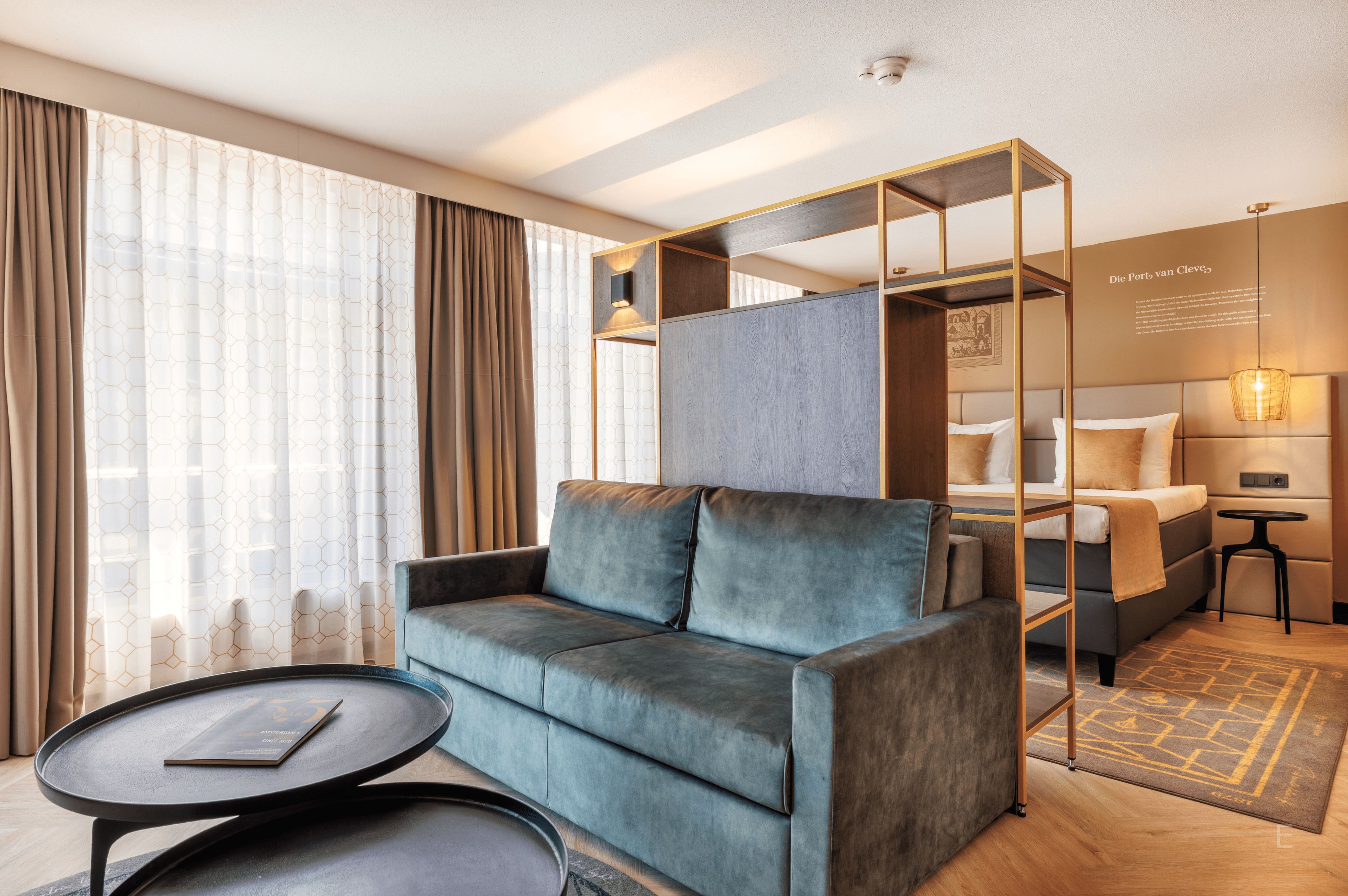 A modern hotel room with a sofa, a small table, a bed with a headboard, and a decorative wall piece.