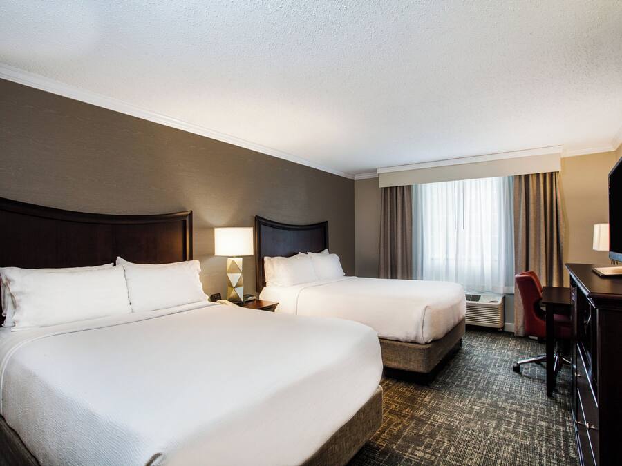 Quarto de hotel com duas camas, uma escrivaninha, uma cadeira, televisão e uma janela com cortinas.