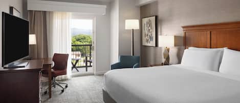 A hotel room with a large bed, a desk with a TV, a chair, and a balcony with a view.