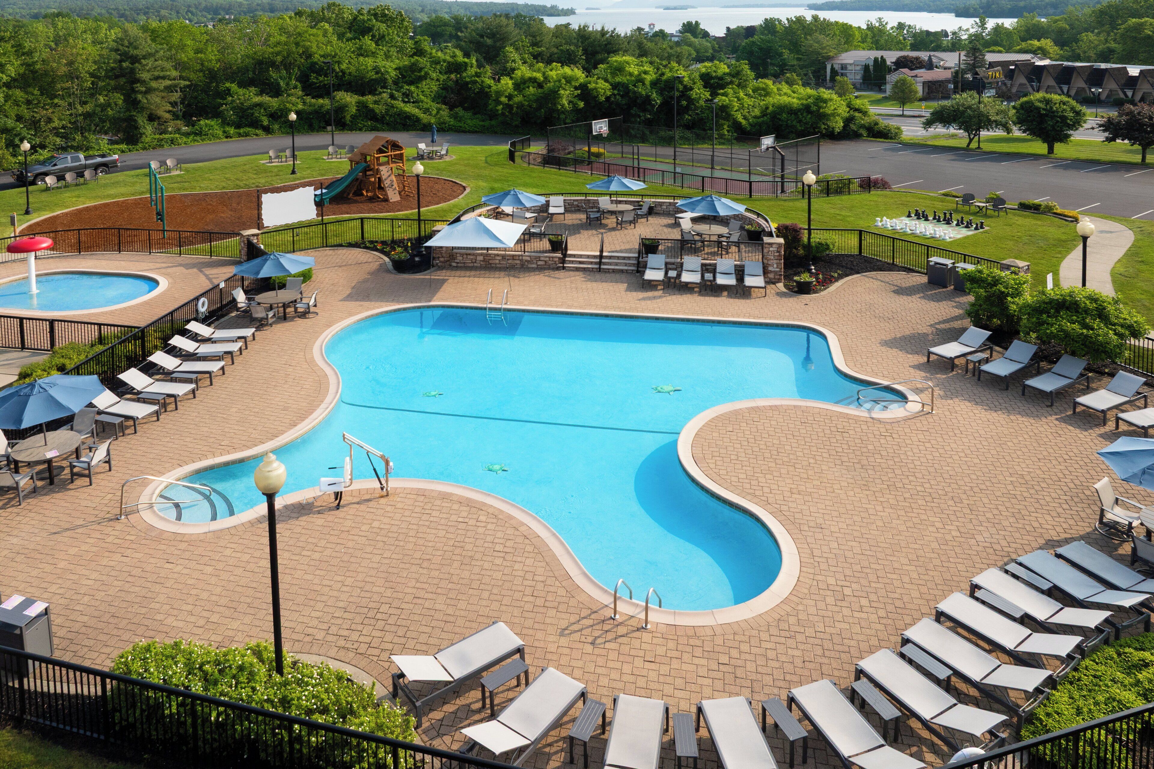 A large outdoor swimming pool with lounge chairs and umbrellas, surrounded by a brick patio and a fenced area with a playground.