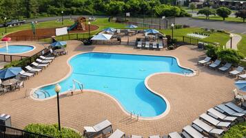 A large outdoor swimming pool with lounge chairs and umbrellas, surrounded by a brick patio and a fenced area with a playground.