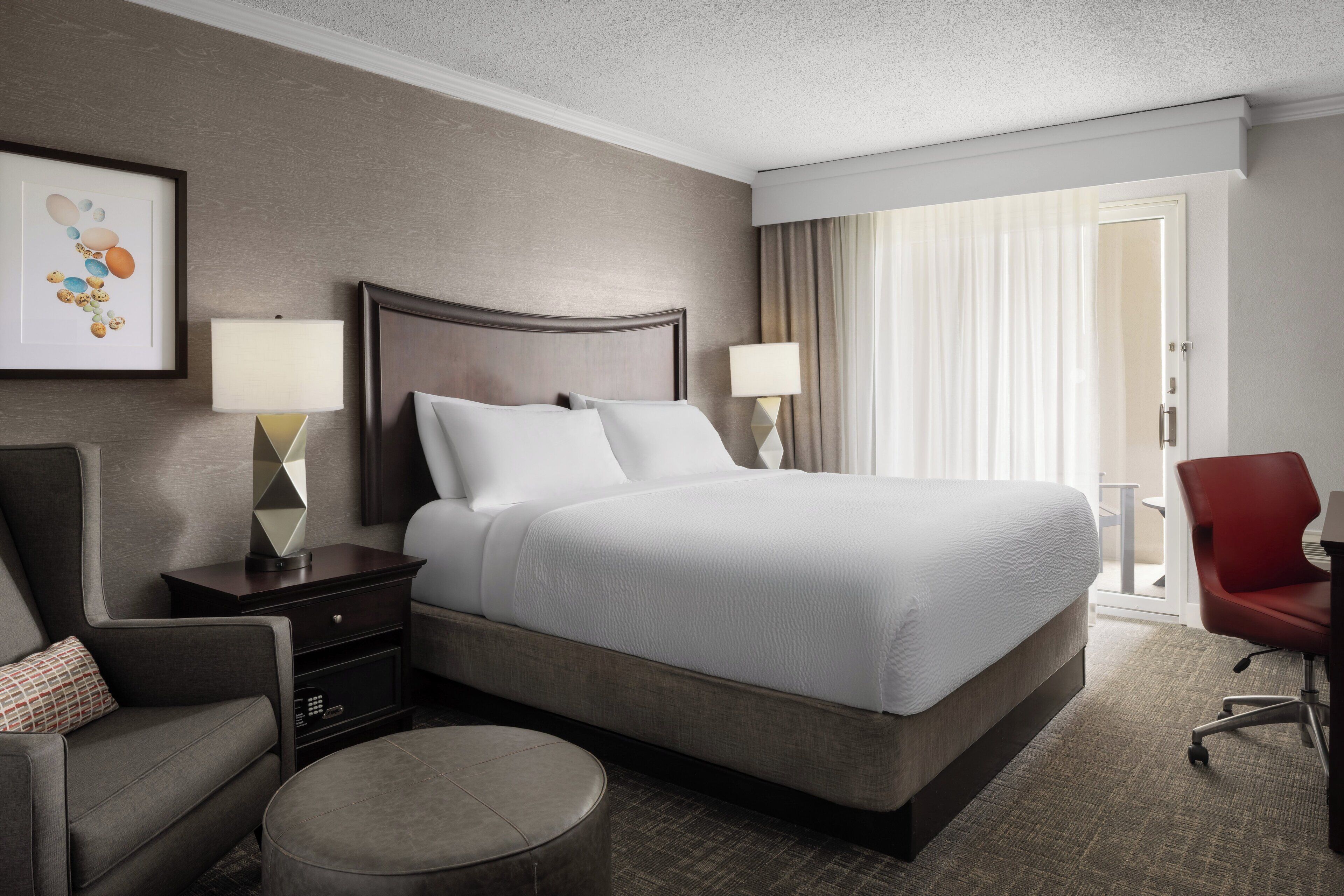 A hotel room with a large bed, two bedside tables with lamps, a chair, and a view of the outdoors through a window with curtains.