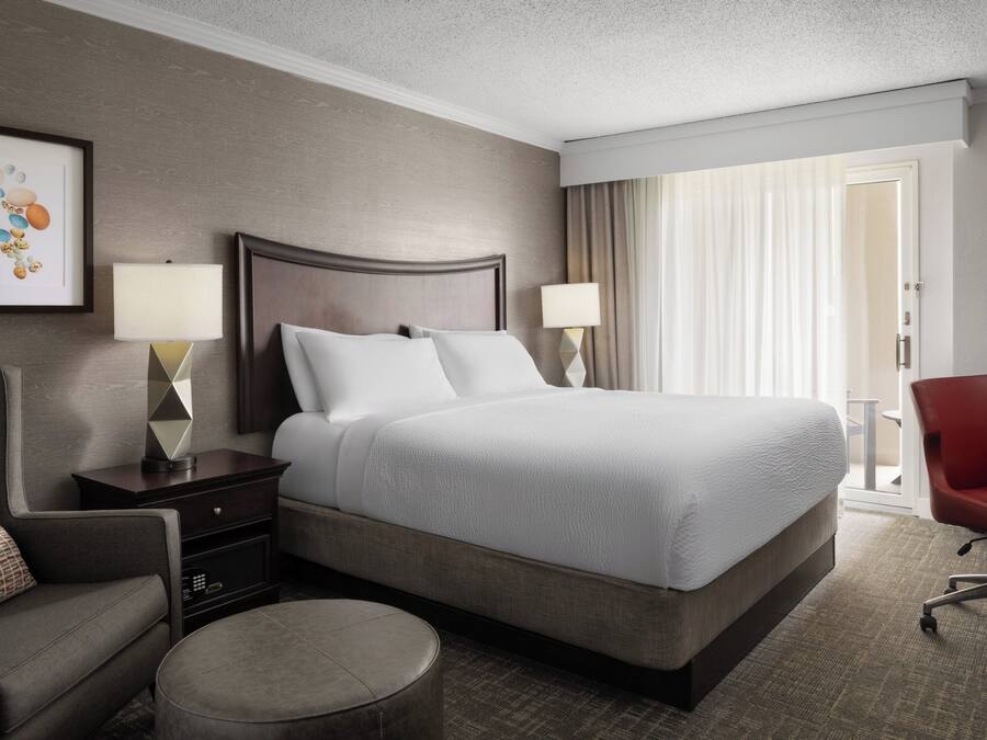 A hotel room with a large bed, two bedside tables with lamps, a chair, and a view of the outdoors through a window with curtains.