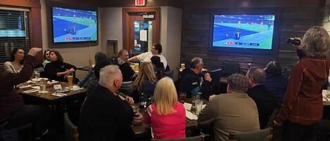 A conference room with multiple screens displaying sports events, tables with drinks, and attendees seated.