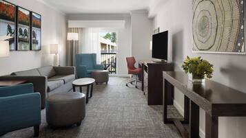 Et moderne hotelværelse med gråt tæppe, en sofa, to lænestole, et sofabord, et fjernsyn, et skrivebord og et stort vindue med udsigt til en bygning og træer.