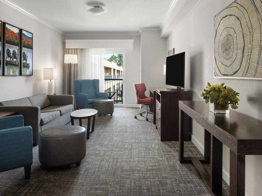 Et moderne hotelværelse med gråt tæppe, en sofa, to lænestole, et sofabord, et fjernsyn, et skrivebord og et stort vindue med udsigt til en bygning og træer.