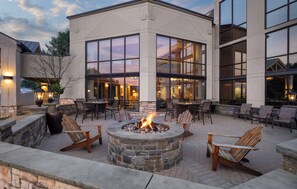 A patio area with a stone fire pit, wooden chairs, and tables, surrounded by a building with large windows and a stone facade. - Holiday Inn Resort Lake George - Adirondack Area by IHG (Lake George)