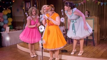 A stage with three individuals in colorful dresses, a microphone stand, and a banner in the background.