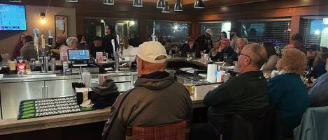 A bar area with multiple patrons seated at tables, a bar counter with a cash register, and overhead lighting.