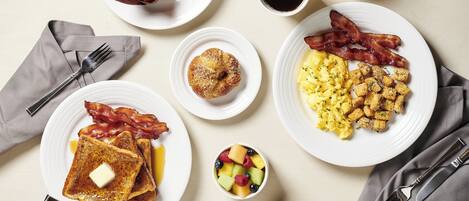 A breakfast setting with toast, eggs, bacon, a muffin, fruit, and beverages on plates.