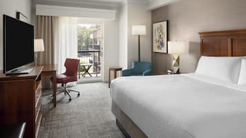 A hotel room with a large bed, a desk with a TV, a chair, a small table, and a balcony with a chair and table.