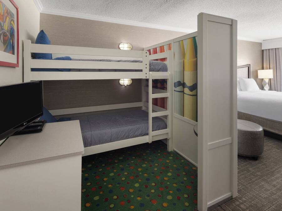 Quarto de hotel com beliche, uma escrivaninha com televisão e uma pequena lixeira.