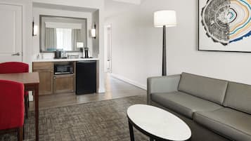 A modern hotel room with a living area, a dining table with red chairs, a gray sofa, a small white table, a kitchen area, and a large abstract artwork on the wall.